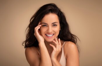 Closeup portrait of sensual beautiful young brunette long-haired woman posing on beige studio background, touching her face and smiling, enjoying her smooth glow face skin after beauty treatment