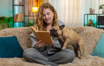 Young relaxed Caucasian redhead woman reads book aloud to pug dog on home sofa with soft voice and warm smile. Pet lies quietly beside her, listening calmly and sharing gentle end-of-day connection.
